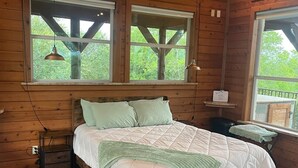 A wooden cabin bedroom with a bed, two windows, a nightstand, and a rug. - The Upper Room | 360° Patio Views & Elevated Hill Country Escape (Buda)