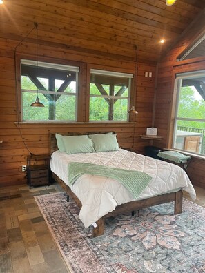 Ein Schlafzimmer in einer Holzhütte mit einem Bett, zwei Fenstern, einem Nachttisch und einem Teppich.