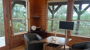 A wooden cabin interior with a sofa, a table, and a large window. - The Upper Room | 360° Patio Views & Elevated Hill Country Escape (Buda)