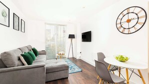 Ett modernt vardagsrum med en grå soffa, ett litet runt bord med stolar, en stor väggklocka och en tv som är monterad på väggen.