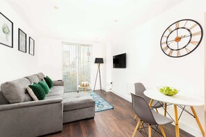 Ein modernes Wohnzimmer mit einem grauen Sofa, einem kleinen runden Tisch mit Stühlen, einer großen Wanduhr und einem an der Wand befestigten Fernseher.