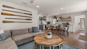 A modern kitchen with white cabinets, a stainless steel refrigerator, and a dining area with wooden chairs and a round table. - A Salty Cru (Panama City Beach)