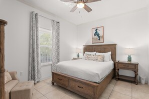 Ein Schlafzimmer mit Bett, Nachttischen, einem Sessel, Deckenventilator und einem Fenster mit Jalousien.