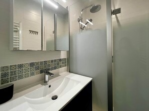 Ein modernes Badezimmer mit einem weißen Waschbecken, einem Spiegel, einer Dusche inklusive Handbrause und einer gefliesten Rückwand.