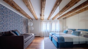 Moderni hotellihuone, jossa on näkyvillä puiset kattopalkit, kuvioitu seinä, sohva, sinisillä tekstiileillä varustettu sänky ja suuri ikkuna, jossa on läpikuultavat verhot.