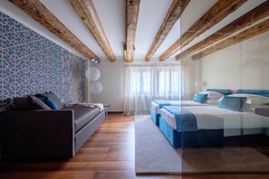 Een moderne hotelkamer met zichtbare houten balken, een patroonmuur, een bank, een bed met blauw beddengoed en een groot raam met dunne gordijnen.