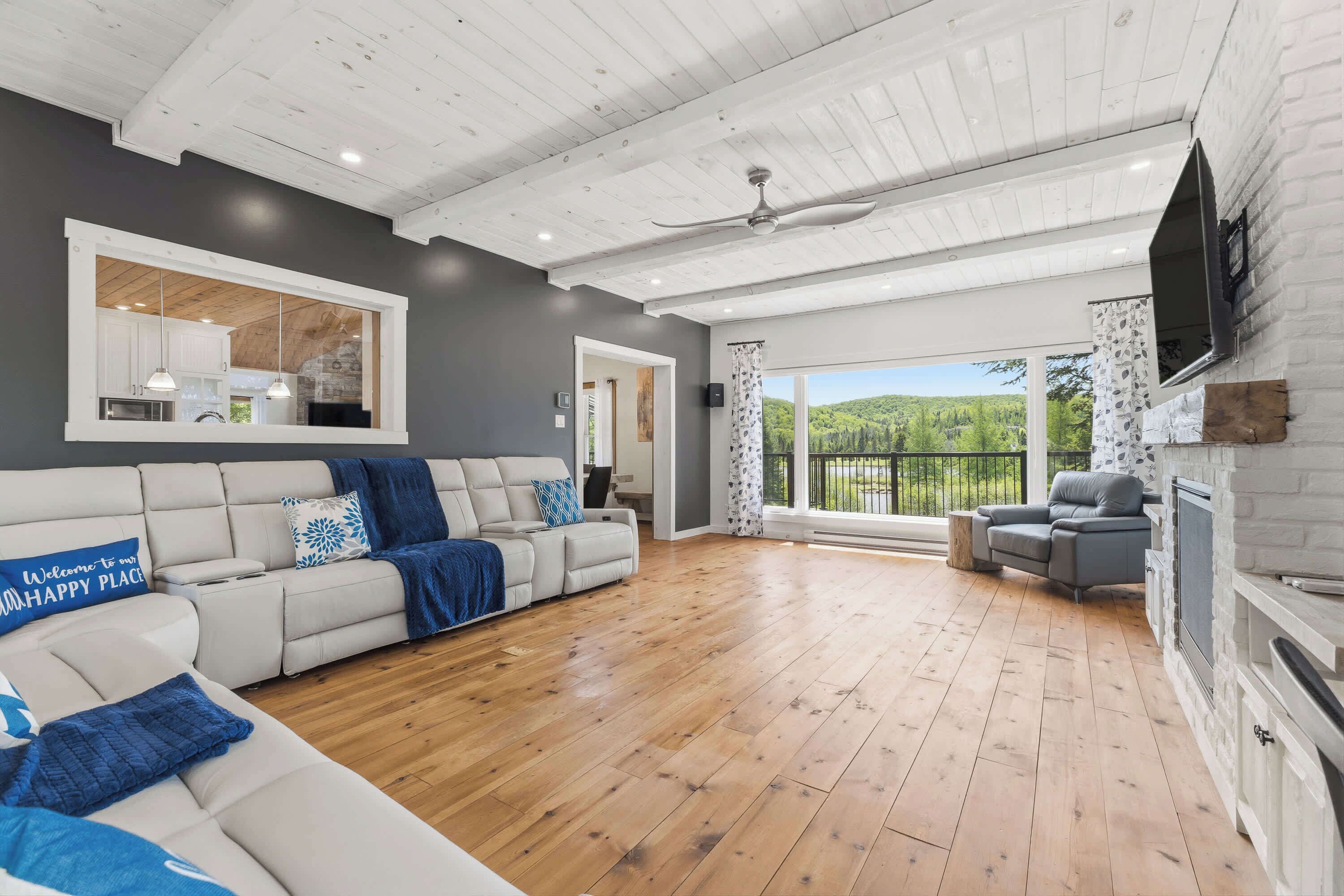 A spacious living area with a wooden floor, a white sofa with blue cushions, a grey sofa, a fireplace, and a large window offering a view of greenery.