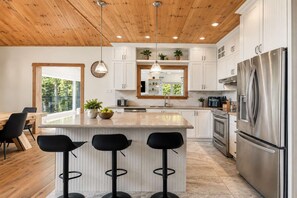 A modern kitchen with a wooden ceiling, white cabinets, a stainless steel refrigerator, and a bar area with stools. - Spa & pool Access - Refuge au fil des saisons (Mont-Blanc)