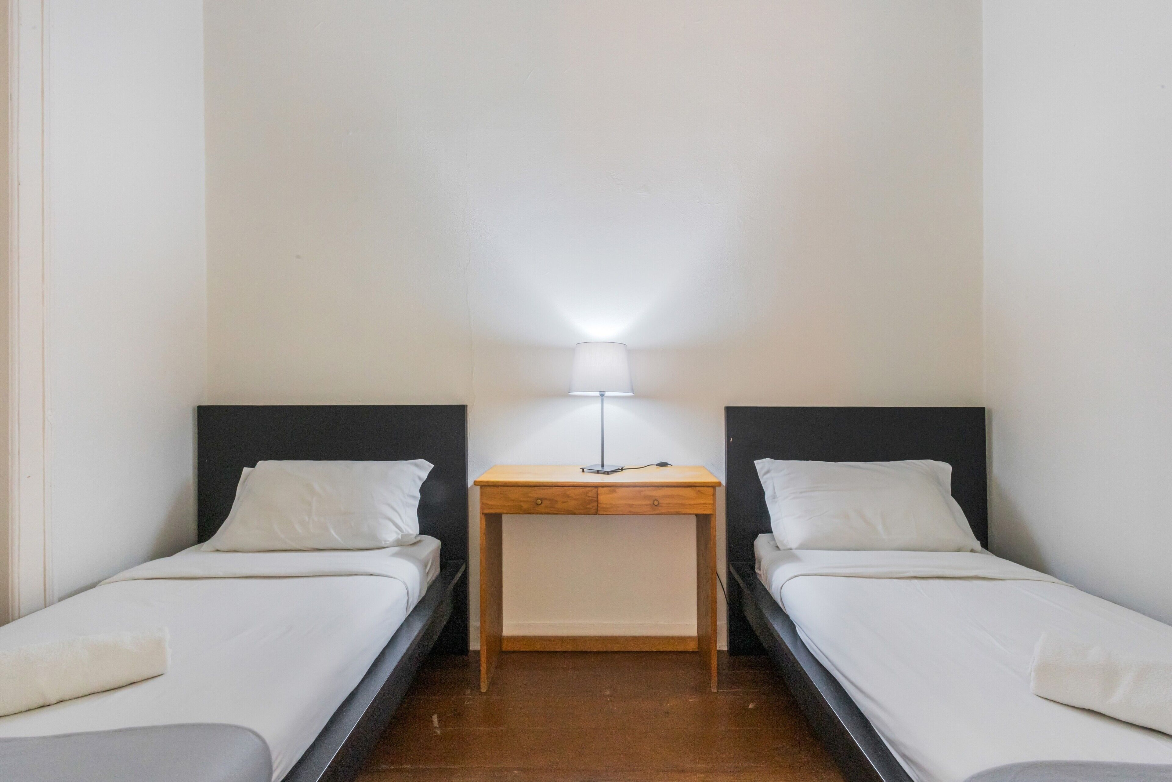 A small, clean room with two single beds, a bedside table with a lamp, and a wooden floor.