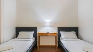 A small, clean room with two single beds, a bedside table with a lamp, and a wooden floor.