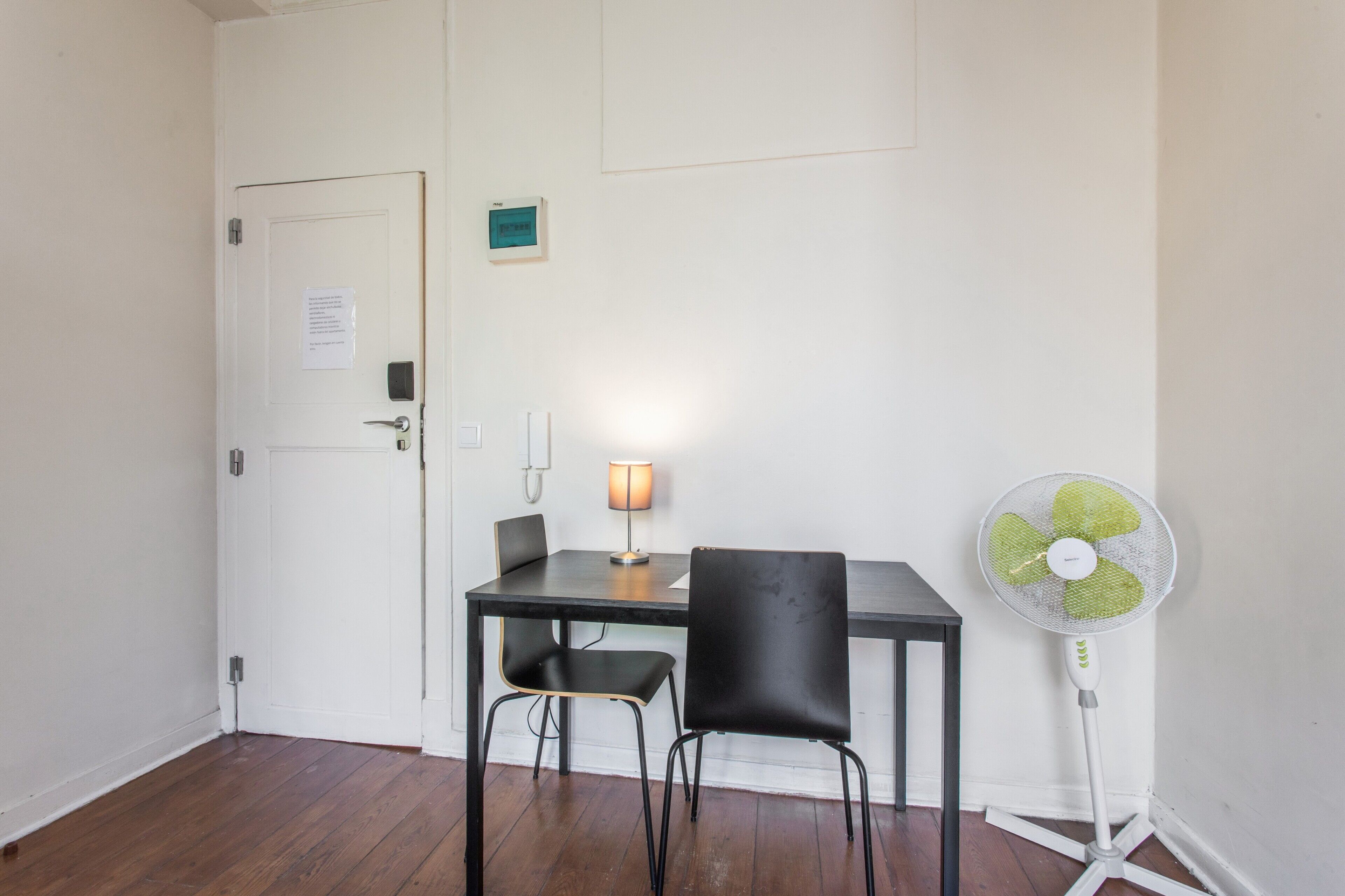Una habitación con piso de madera, una puerta blanca, una mesa con una silla, un ventilador de pie y un teléfono montado en la pared.