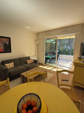 A living room with a sofa, a dining table with chairs, a TV stand, and a sliding glass door leading to an outdoor patio. - T2 luxury residence in a wooded park with 2 private pools and seafront (Saint-Raphaël)