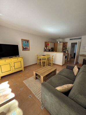 A living room with a TV, yellow cabinets, a dining table with chairs, a grey sofa, and a kitchen in the background. - T2 luxury residence in a wooded park with 2 private pools and seafront (Saint-Raphaël)