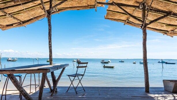 Una terraza de madera con mesa y sillas, con vista a aguas tranquilas y varias embarcaciones.