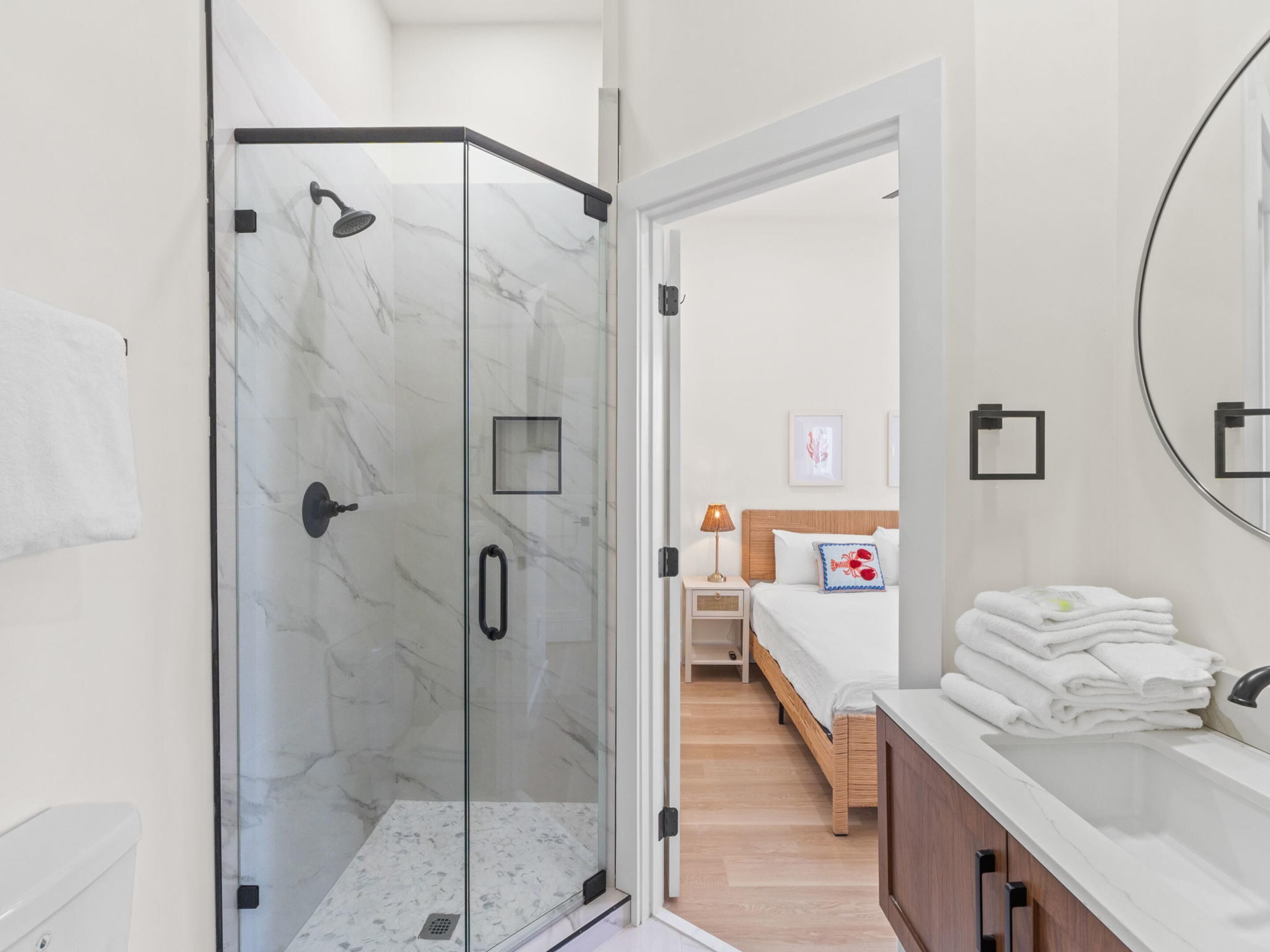 Een moderne badkamer met een douchecabine van glas, een houten vloer, een witte wastafel met spiegel en een netjes opgemaakt bed met kussen.