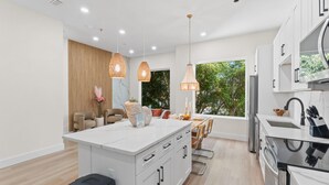 Uma cozinha moderna com armários brancos, uma ilha central, luminárias pendentes e vista para o verde lá fora.