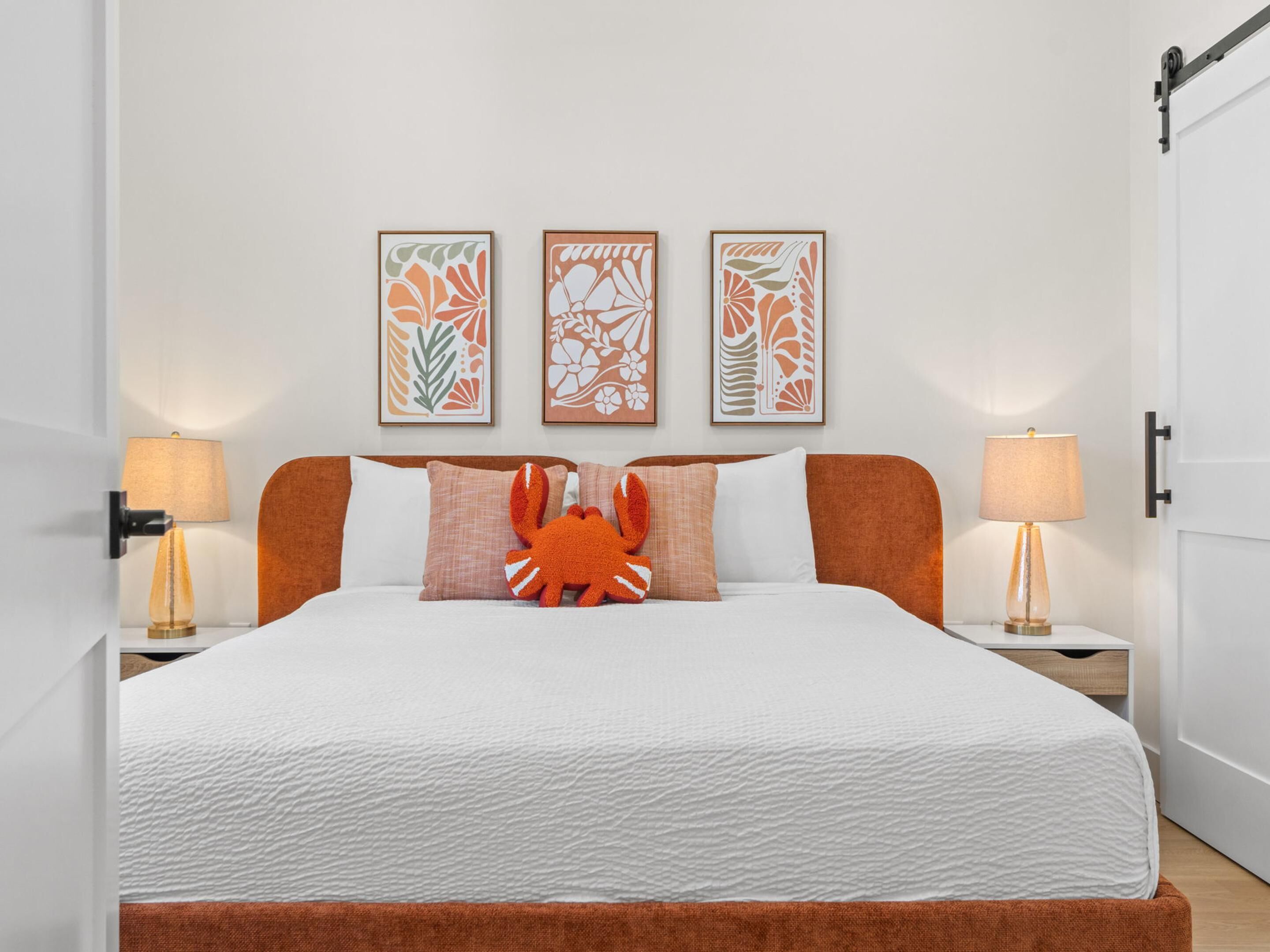 Una cama bien hecha con edredón blanco, cabecera marrón y un cojín decorativo con forma de cangrejo. Encima de la cama hay tres obras de arte enmarcadas, y dos lámparas de noche a cada lado.