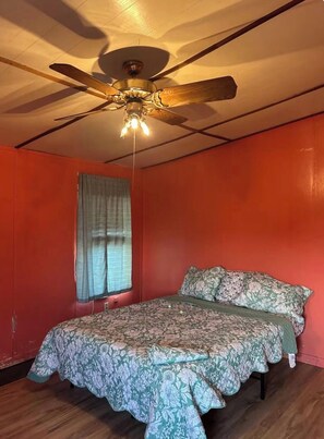 Une chambre à coucher avec un ventilateur de plafond, un lit recouvert d’une literie à motifs floraux et une fenêtre avec des rideaux.