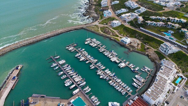 An aerial view of a marina with numerous boats docked, surrounded by residential buildings and a rocky coastline. - Skal to 8 on Serifos (Langebaan)