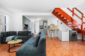 Un salon comprenant un canapé, une table basse et une cheminée. On y trouve également un coin cuisine avec une table à manger et des chaises, ainsi qu’un espace buanderie avec un lave-linge. Un escalier en bois est visible.