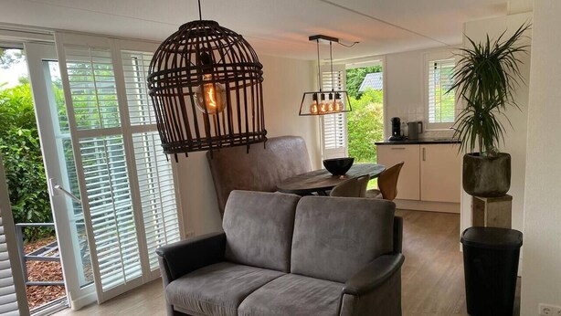 A modern living room with a grey sofa, a hanging light fixture, and a view of the outdoors through large windows. - Eendje 2 | Luxe recreatiewoning met hottub (Ermelo)