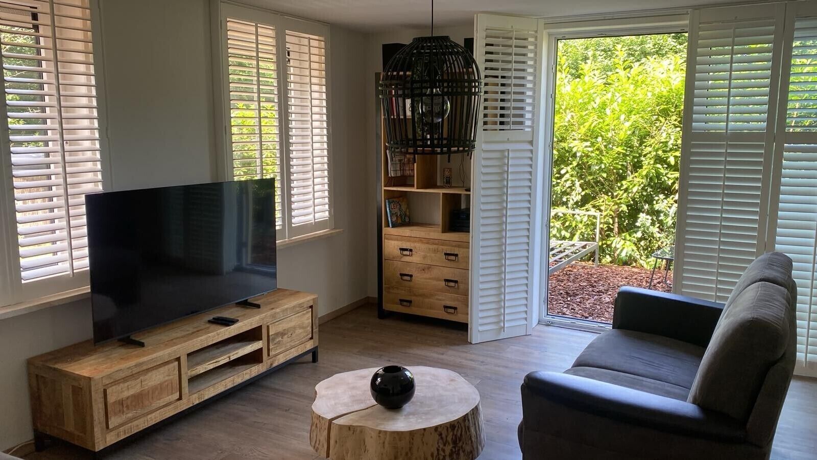 A living room with a flat-screen TV on a wooden TV stand, a grey sofa, a round coffee table with a black vase, and a bookshelf with open white shutters.