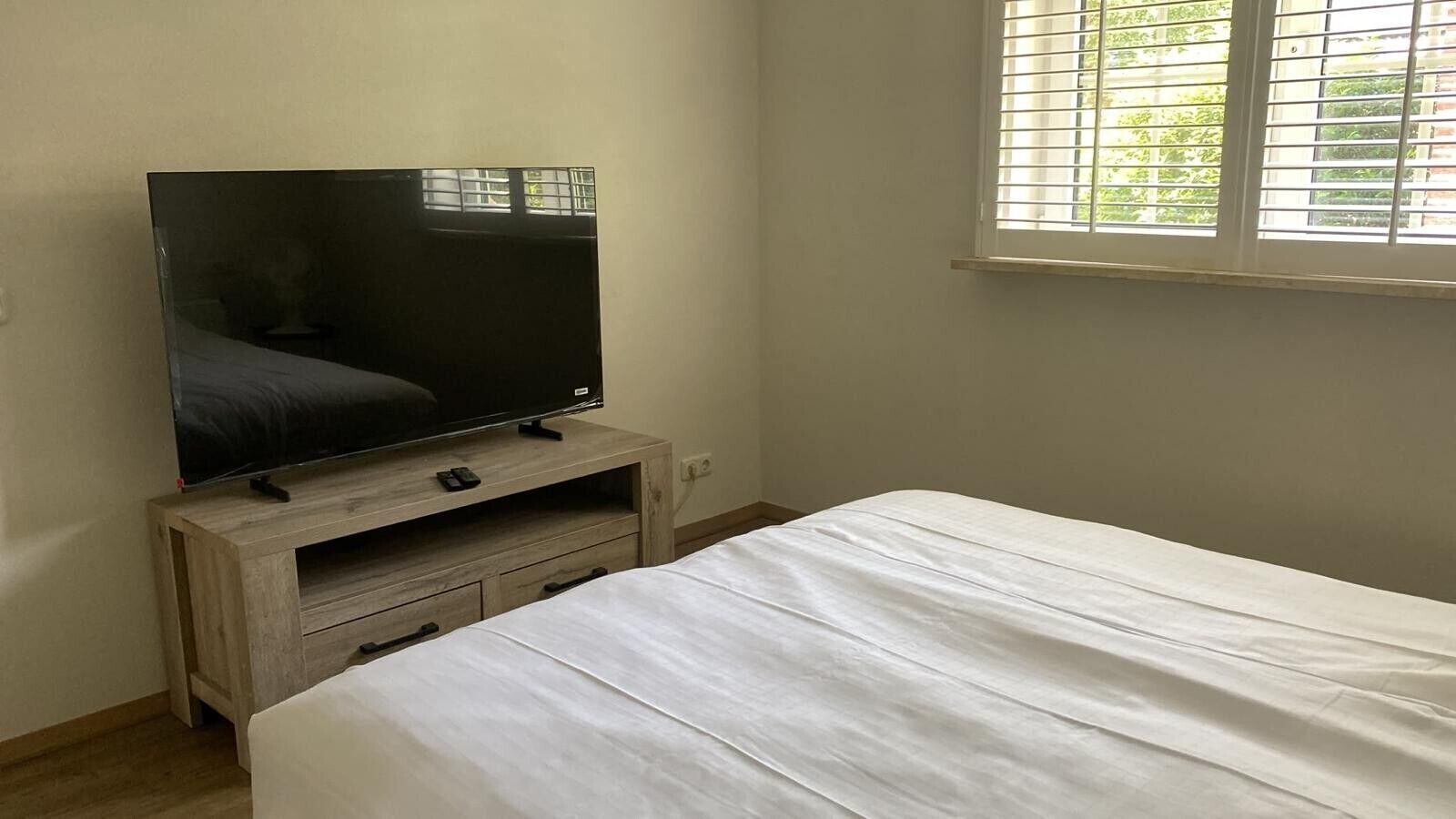 A bedroom with a bed, a TV on a wooden stand, and a window with blinds.