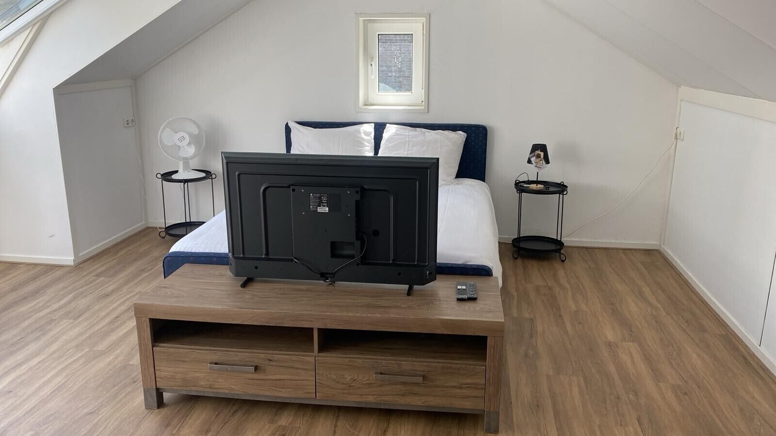 A bedroom with a bed, a TV on a wooden stand, a bedside table with a lamp, and a small table with a remote control.