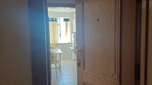 Un pasillo que conduce a una habitación con puerta de madera, un comedor con mesa y sillas, y un ventanal con cortinas.