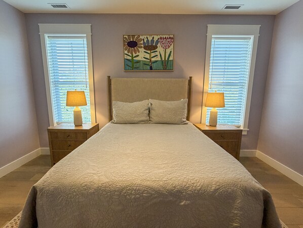 A bedroom with a bed, two bedside tables with lamps, a painting on the wall, and windows with blinds.