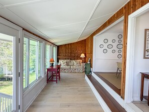 A room with a wooden paneled wall, a sofa, a table with a lamp, and a view through large windows. - DC56: Silver Queen| Centrally Located | New Decks (Ocracoke)