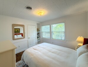 A bedroom with a bed, a dresser, a nightstand, a window with blinds, a door, and a ceiling vent. - DC56: Silver Queen| Centrally Located | New Decks (Ocracoke)