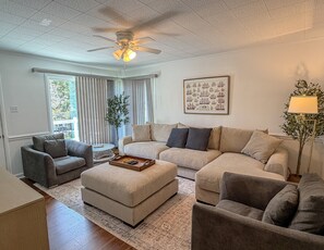 A living room with a sectional sofa, a grey armchair, a coffee table with remote controls, and a ceiling fan. - DC56: Silver Queen| Centrally Located | New Decks (Ocracoke)