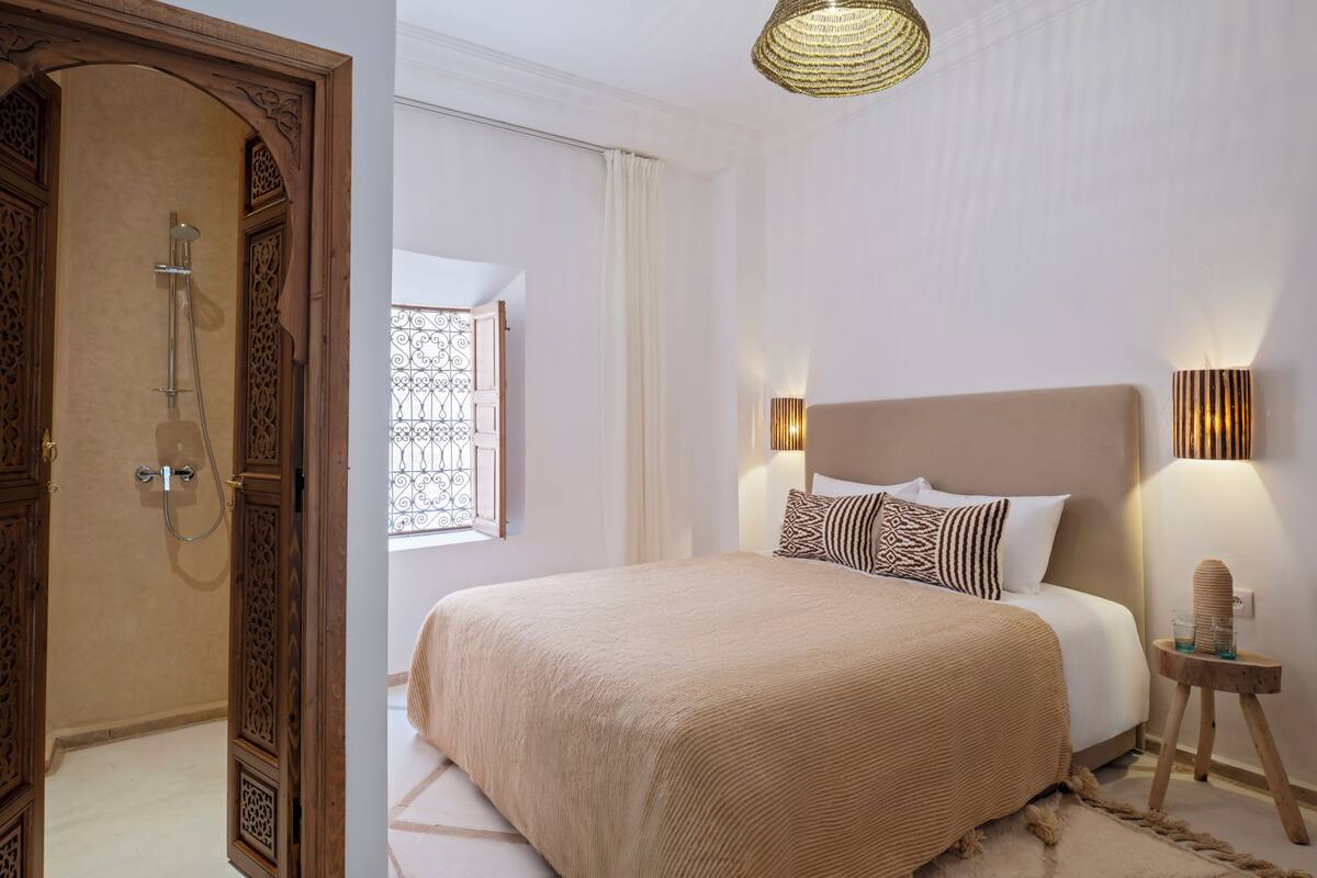 A bedroom with a large bed, a wooden door, a window with a metal grille, and a wall-mounted lamp.