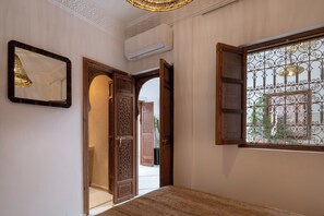 A room with a bed, an open door leading to a hallway, a wall-mounted air conditioner, and a window with decorative iron bars.