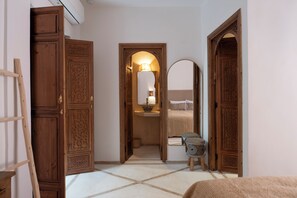 Um corredor com portas de madeira, uma cama com almofadas, um banquinho e um abajur.