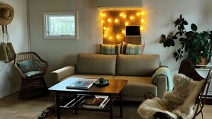Ein gemütliches Wohnzimmer mit einer Couch, einem Korbsessel, einem Couchtisch mit Zeitschriften und einem an der Wand montierten Fernseher.