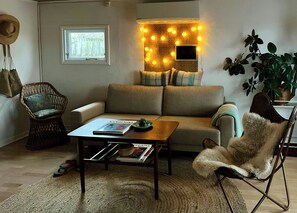 Uma sala de estar aconchegante com um sofá, uma poltrona de vime, uma mesa de centro com revistas e uma TV fixada na parede.