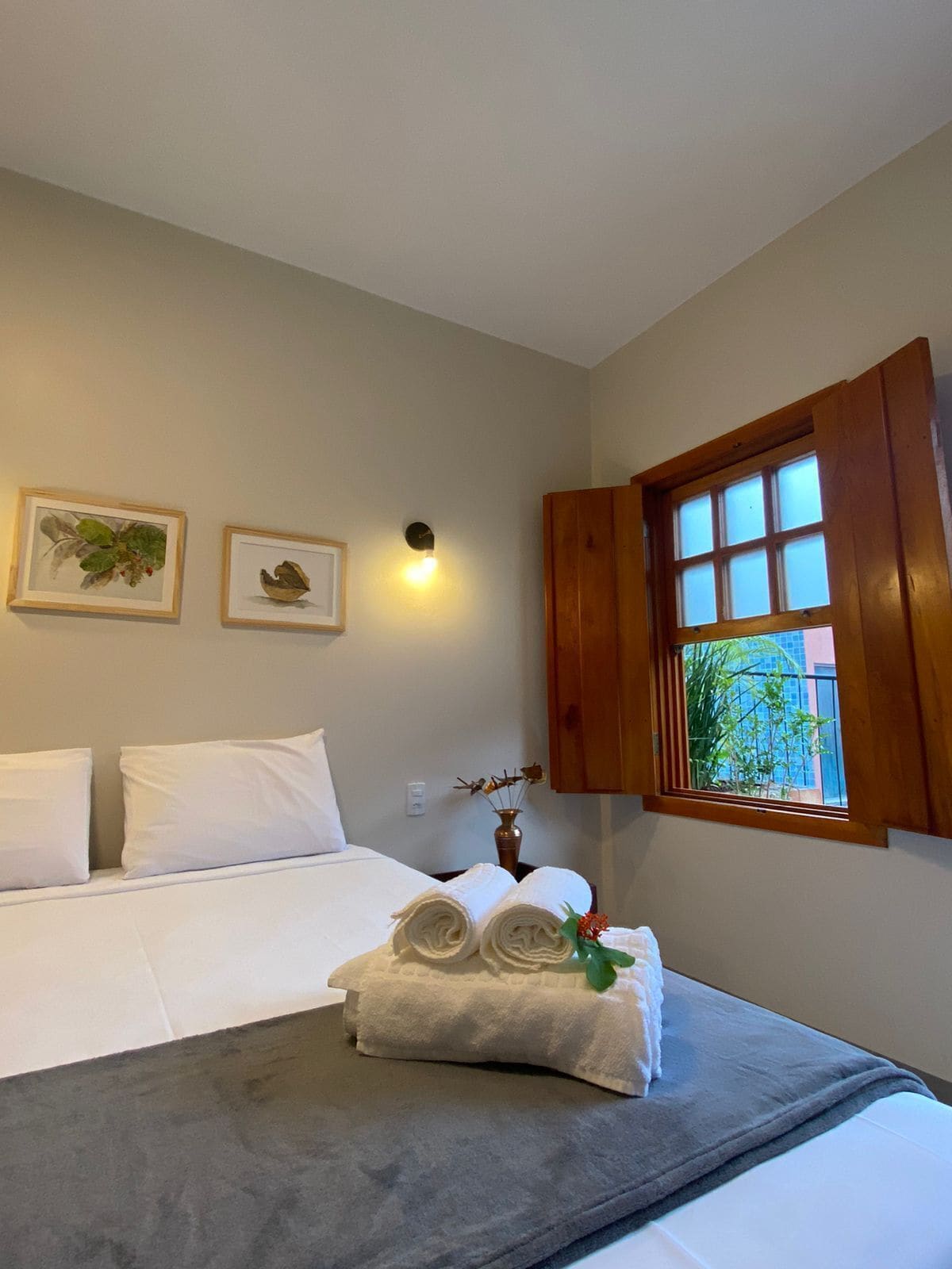 Ein ordentlich gemachtes Bett mit weißen Bettwäsche, einer grauen Tagesdecke und einer Vase mit Blumen darauf. An der Wand hängen zwei gerahmte Bilder, eine Wandleuchte und ein Fenster mit Holzjalousien.