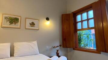 Ein ordentlich gemachtes Bett mit weißen Bettwäsche, einer grauen Tagesdecke und einer Vase mit Blumen darauf. An der Wand hängen zwei gerahmte Bilder, eine Wandleuchte und ein Fenster mit Holzjalousien.