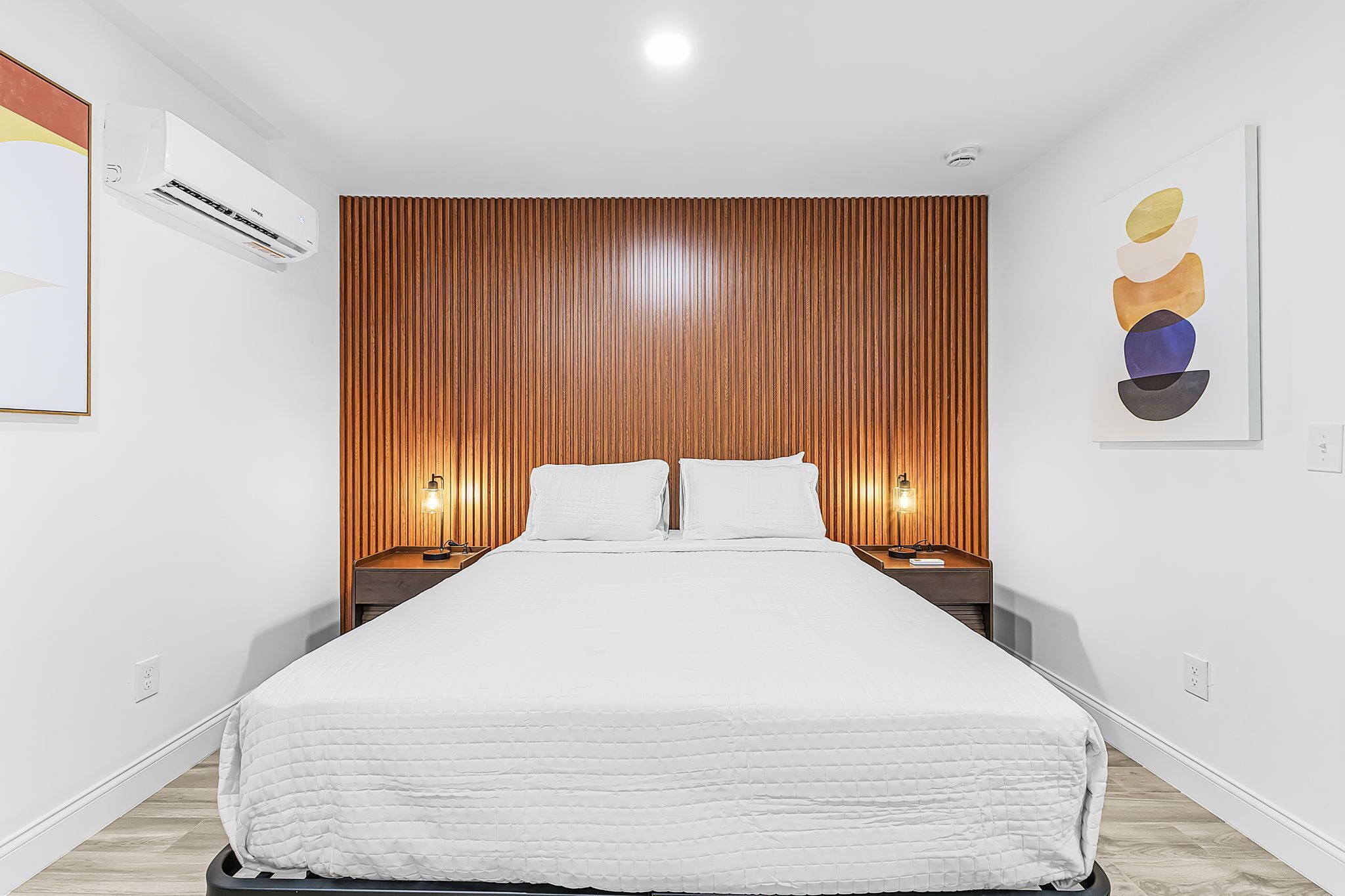 A neatly made bed in a modern room with a wooden panel wall, a wall-mounted air conditioner, and a single artwork on the wall.
