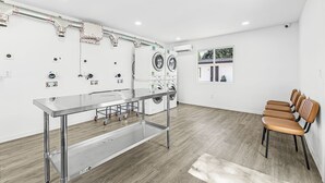 A modern laundry room with stainless steel appliances, a metal table, and wooden chairs. - Veras Inn & Suites (Fort Myers)