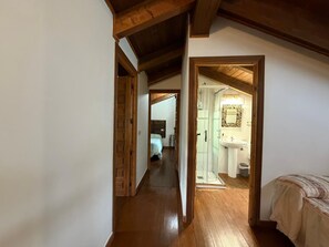 A hallway leading to a bedroom with a wooden ceiling, a visible bathroom with a sink and mirror, and a bed with a patterned bedspread.
