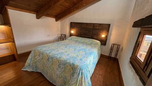 A bedroom with a wooden ceiling, a bed with a patterned bedspread, two bedside lamps, and a window with a view.