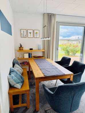 A dining area with a wooden table, benches, and chairs, a small shelf with decorative items, and a view of a garden through a glass door.