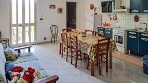 Una zona de menjador amb taula i cadires, un sofà amb coixí floral, i una cuina amb armaris i electrodomèstics.