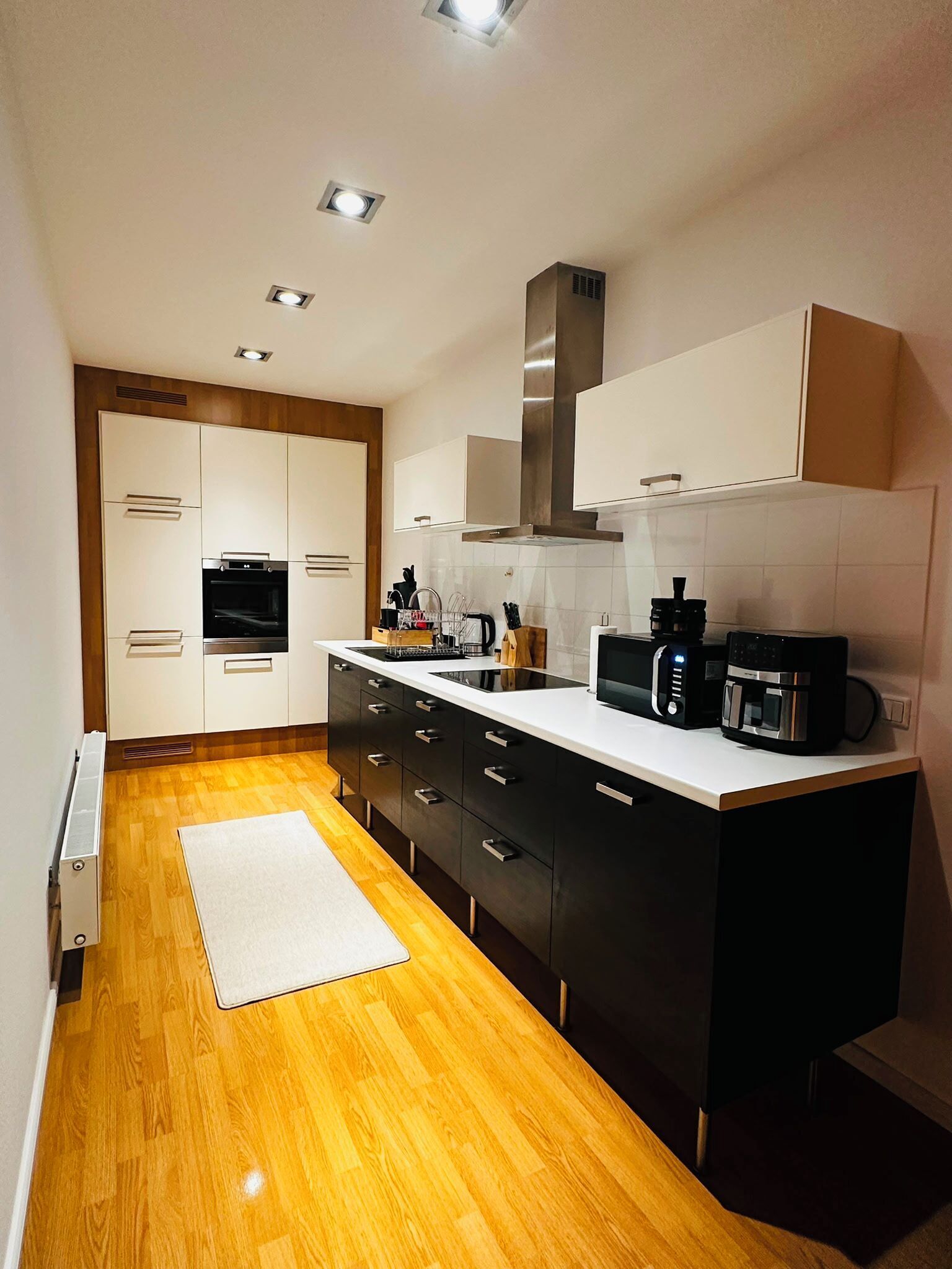 A modern kitchen with wooden flooring, white cabinets, a built-in oven, and a coffee maker.