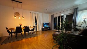 Ein modernes Wohnzimmer mit Holzboden, einer Essecke mit Tisch und Stühlen, einer Couch, einem Fernseher und einer Zimmerpflanze.