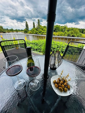 Una mesa con dos copas de vino, una botella y un plato de brochetas, ubicada en un balcón con vista a la vegetación y un río.