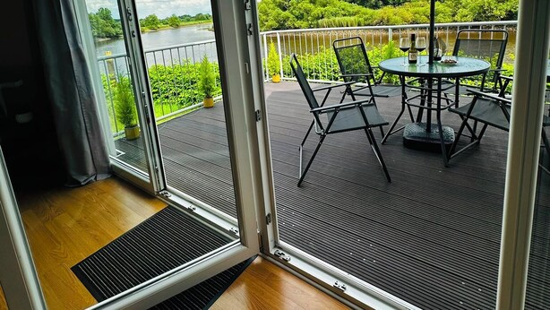 Ein Zimmer mit einer Schiebeglastür, die auf eine Terrasse mit Tisch und Stühlen sowie einen großen Sonnenschirm führt. Blick auf einen See und grüne Vegetation.